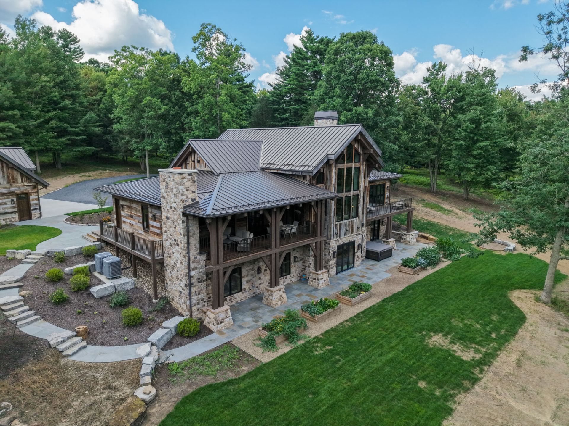 Aerial view of a custom stone-and-timber KB Builders home with covered porch, landscaped patio, and wooded lot in Central Pennsylvania