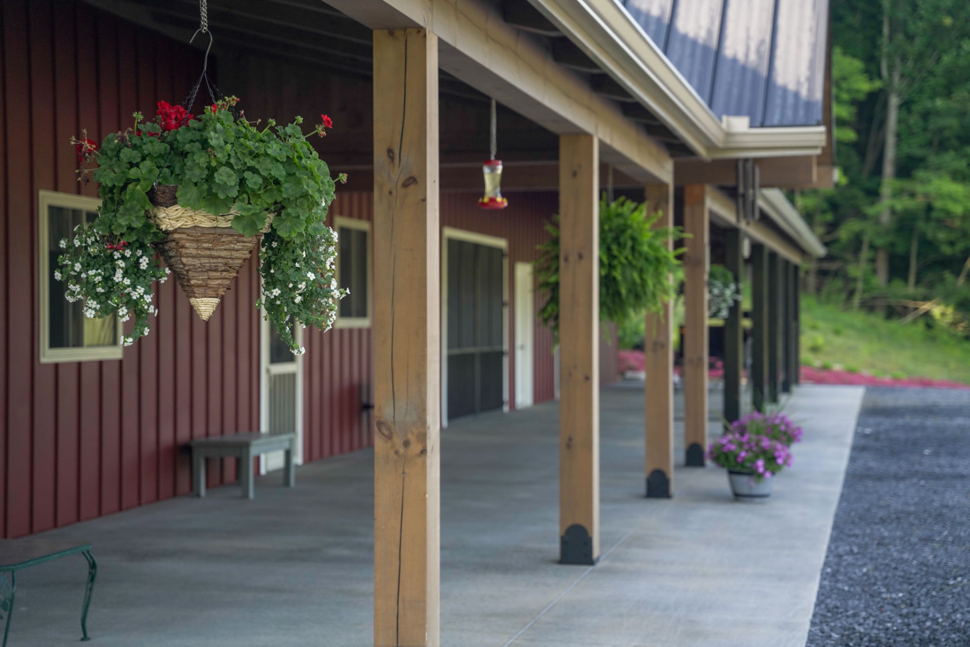 Peachy Residence — covered porch with timber posts, hanging flower baskets, and bench along barn-red siding — KB Builders, McVeytown, PA